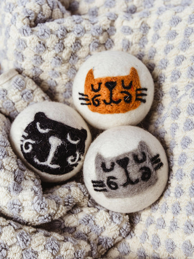 Trio of wool dryer balls with playful cat faces in orange, black, and gray felt, arranged on a textured cream and gray  bath towel for a cozy, handmade look.