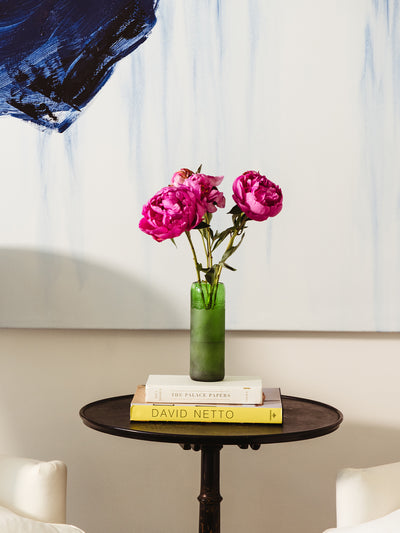 A small black round side table holds a green glass vase with bright pink peonies, resting atop two stacked books titled The Palace Papers and David Netto. A large abstract painting with bold blue brushstrokes hangs on the wall behind the arrangement, adding a modern, artistic flair.