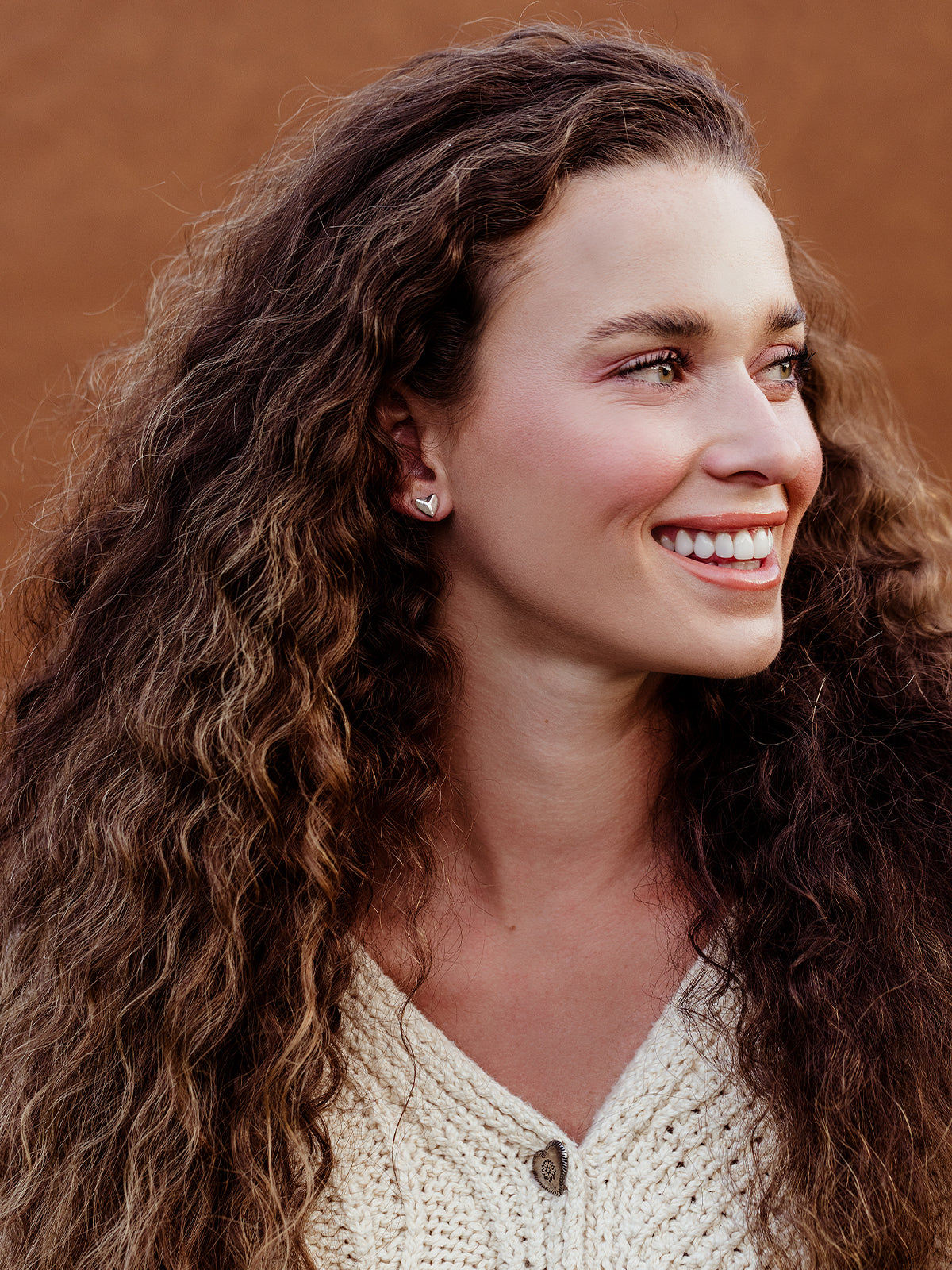Smiling woman with curly hair wearing silver heart stud earrings, minimalist ethical jewelry styled with a cream knit sweater.