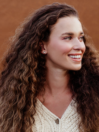 Smiling woman with curly hair wearing silver heart stud earrings, minimalist ethical jewelry styled with a cream knit sweater.