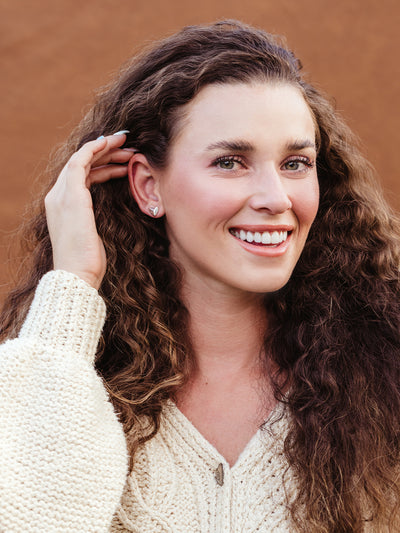 Woman adjusting her hair while wearing dainty silver heart stud earrings, handcrafted ethical jewelry styled with cozy knit fashion.