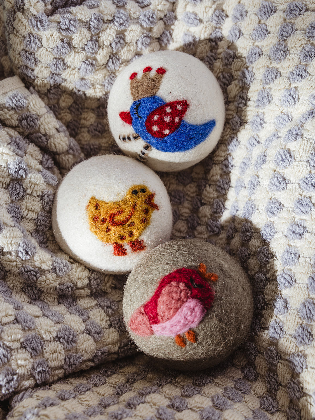 Set of three wool dryer balls with colorful hand-felted bird designs, including a blue bird with a crown, a yellow chick, and a red bird on a soft woven cream and gray textile background.