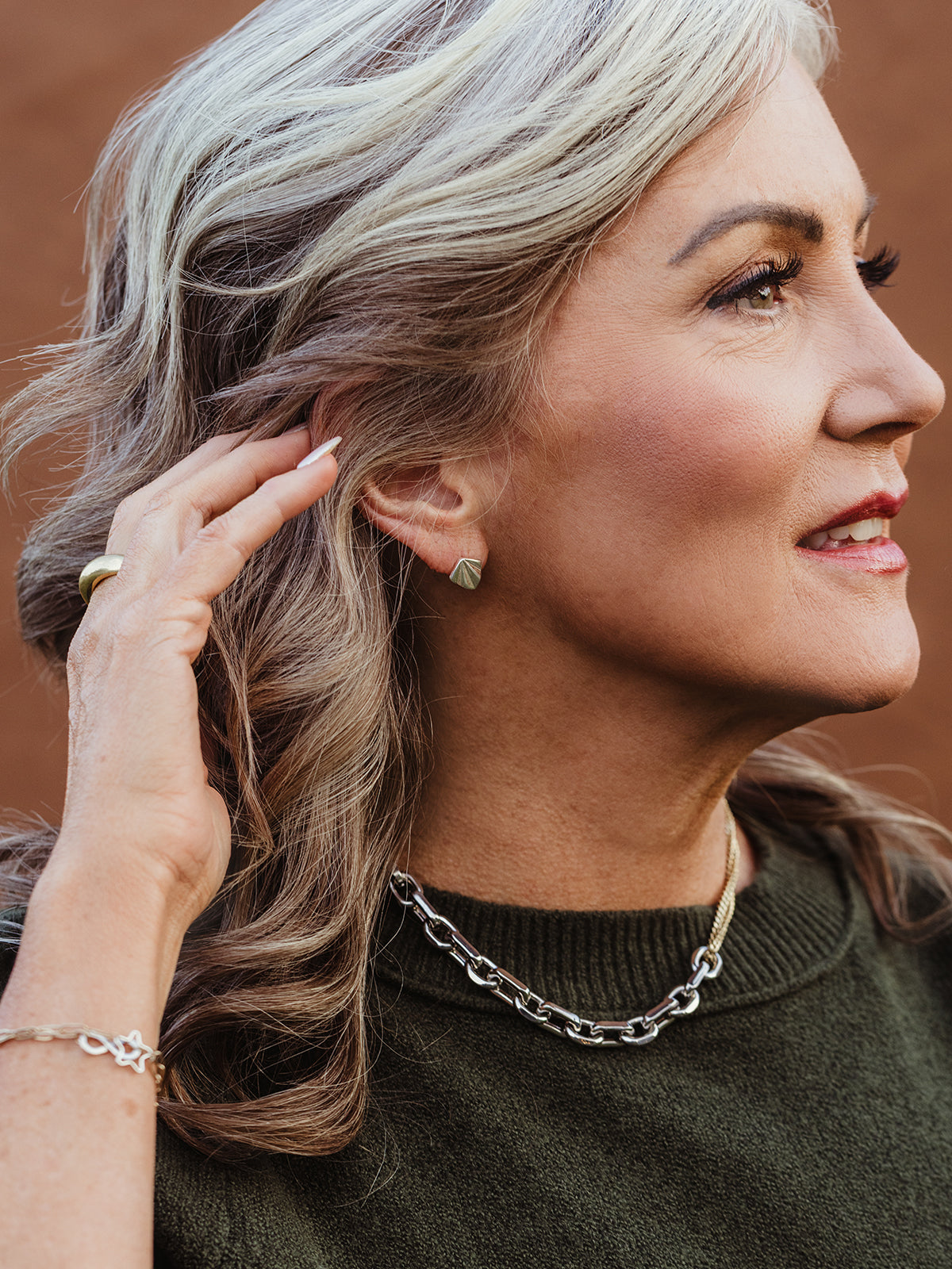Side profile of woman adjusting her hair while wearing silver stud earrings, chunky chain necklace, and artisan bracelet, ethical jewelry styling inspiration.