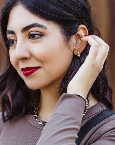 Close-up of woman wearing chunky gold hoop earrings, silver chain necklace, and metallic beaded bracelet – modern ethical jewelry style.