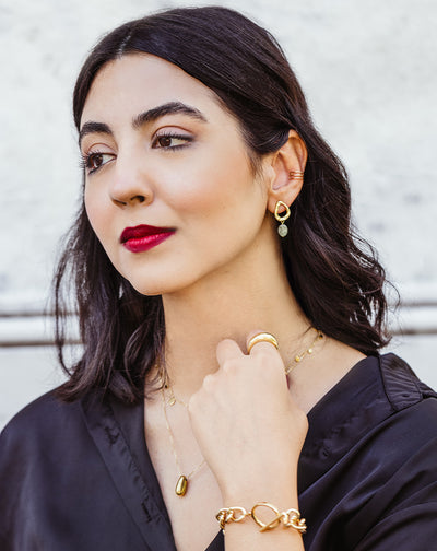 Woman in black blouse wearing gold teardrop dangle earrings, layered pendant necklaces, bold gold ring, and chunky chain bracelet – artisan handmade jewelry collection.