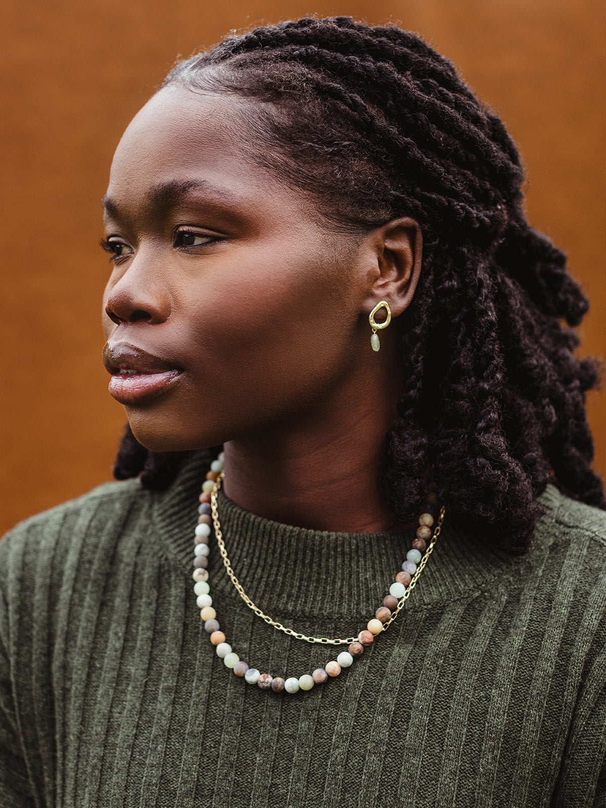 Woman in green ribbed sweater wearing gold drop earrings with green gemstone and layered necklaces—one gold chain and one natural stone beaded necklace.
