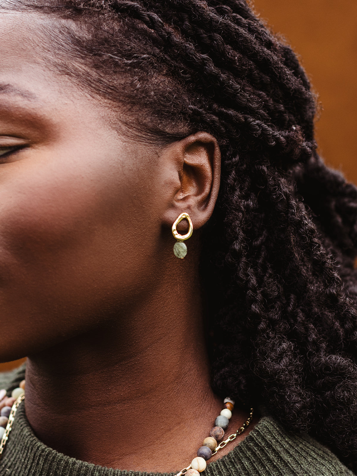 Close-up of gold drop earring with green gemstone worn by woman, styled with layered stone beaded necklace and gold chain.