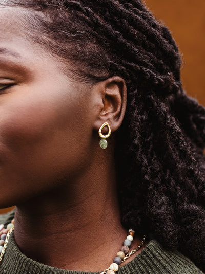 Close-up of gold drop earring with green gemstone worn by woman, styled with layered stone beaded necklace and gold chain.