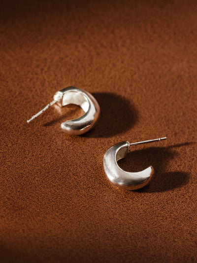 Chunky crescent-shaped sterling silver hoop earrings on leather-textured surface – bold artisan jewelry design.