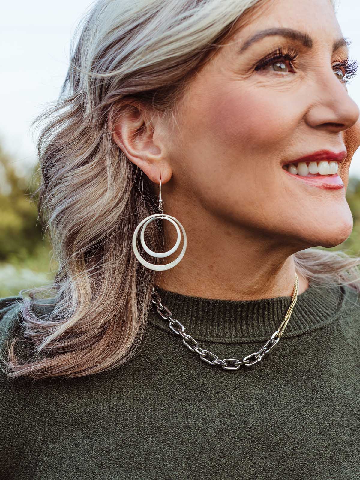 Woman wearing handcrafted double hoop silver earrings and a chunky chain necklace with a dark green sweater — ethically made minimalist jewelry.
