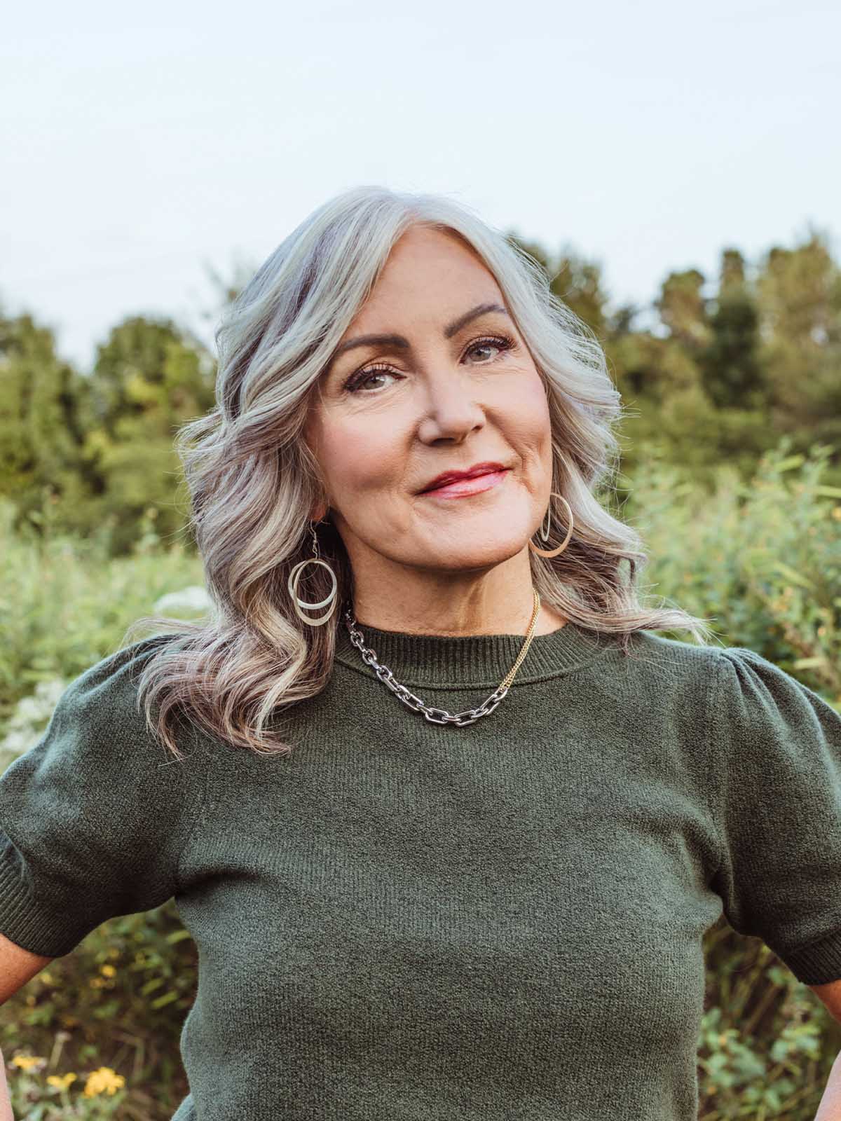 Smiling woman wearing double hoop silver earrings and layered chain necklace with dark green sweater — ethical handcrafted jewelry for everyday style.