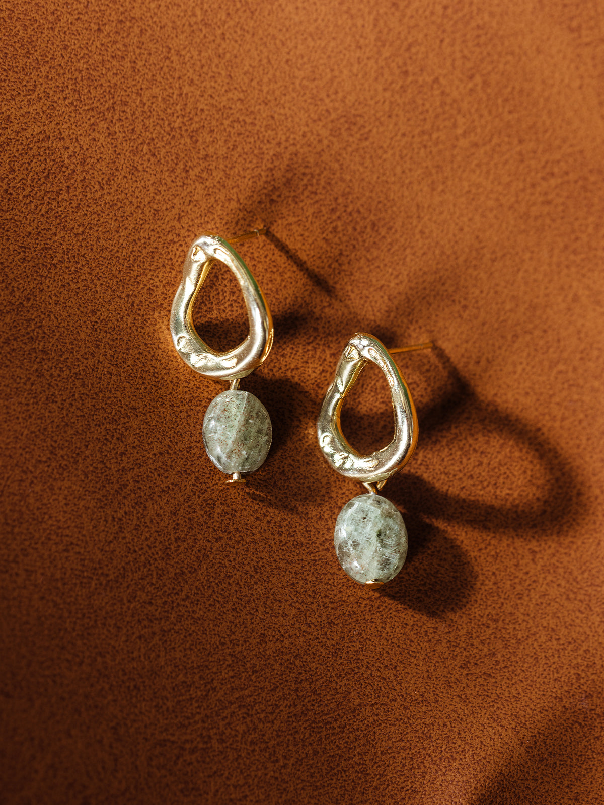Pair of artisan-made gold drop earrings with organic open teardrop design and oval gray stone accents, styled on a warm brown background — ethically handcrafted jewelry.