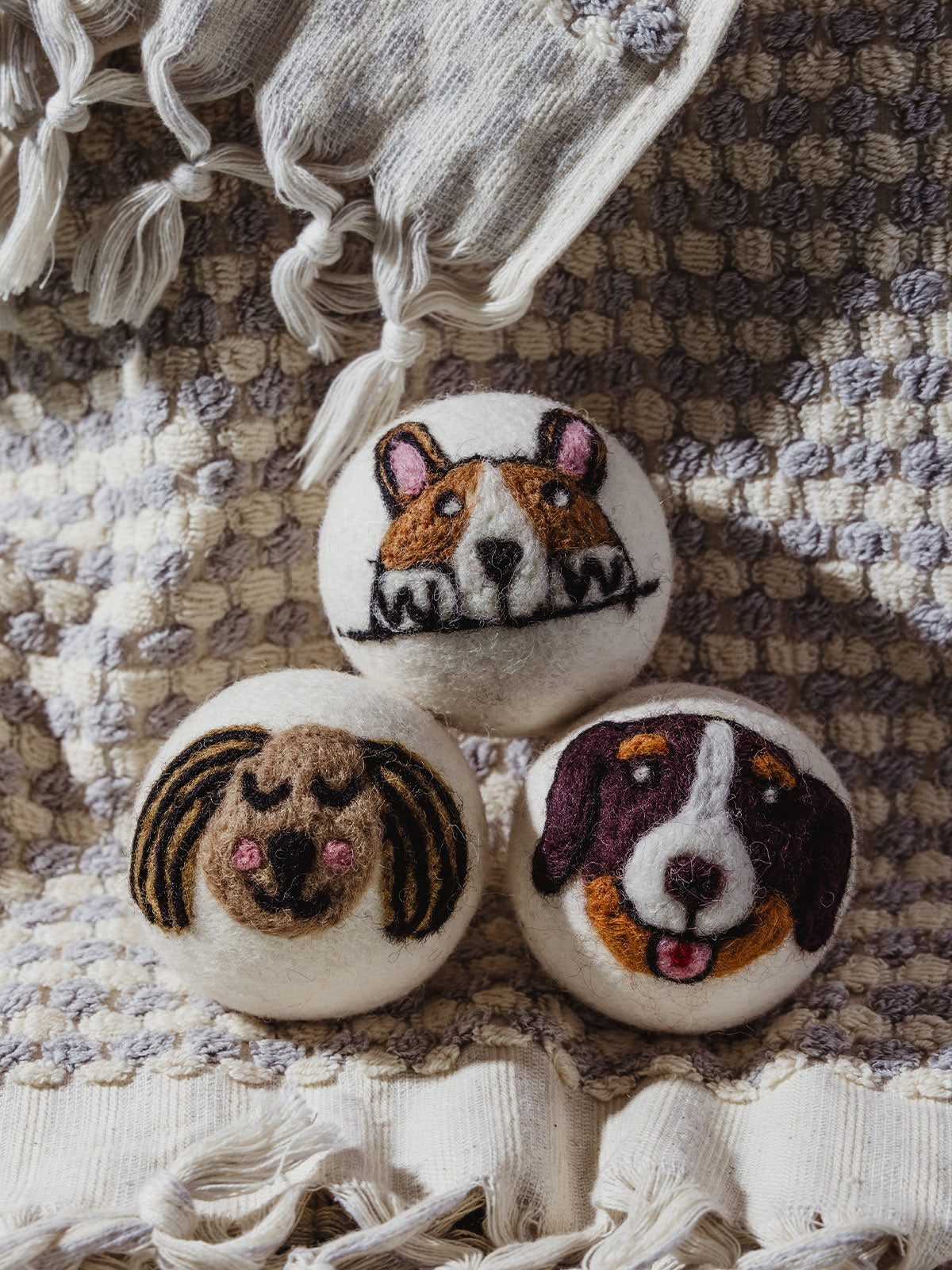 Three wool dryer balls featuring hand-felted dog faces – a brown and white corgi, a brown floppy-eared pup, and a dark brown dog with a pink tongue – stacked on a textured cream and gray blanket.