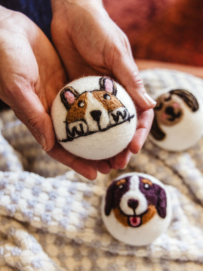 lose-up of hands holding a white wool dryer ball featuring a hand-felted dog face, with two more illustrated dog dryer balls resting on a soft, knit blanket in the background.