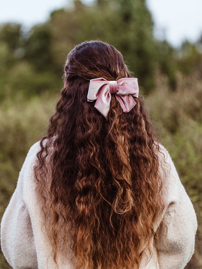 Woman with long wavy brown hair wearing an oversized blush pink velvet hair bow – handmade accessory styled outdoors for an elegant everyday look.