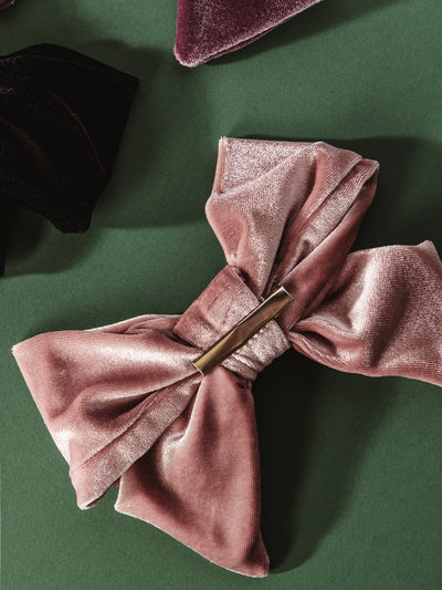 Elegant dusty rose velvet bow hair clip with a gold bar center clasp, styled on a deep green background. Perfect for holiday accessories, sustainable gifting, and ethically made fashion from Joffa.