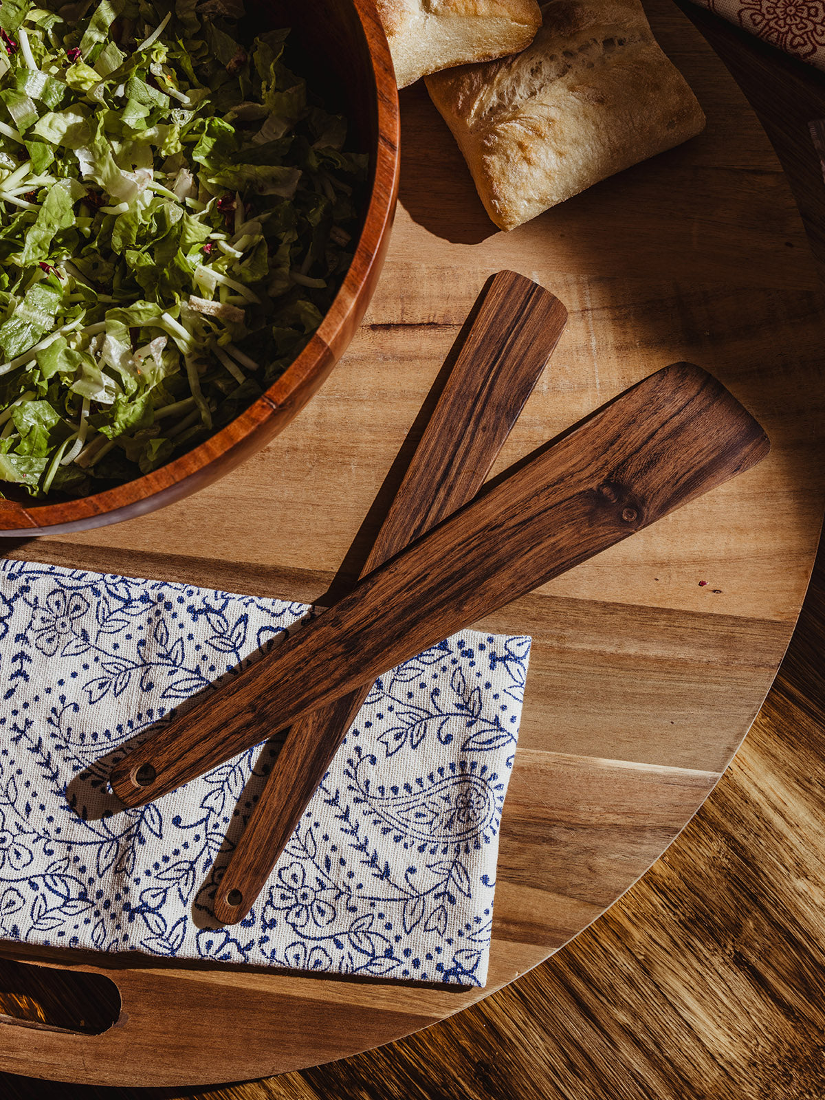  Flat-lay of two wooden salad serving utensils resting on a folded blue floral napkin beside a wooden bowl filled with lettuce and ciabatta bread.