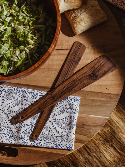  Flat-lay of two wooden salad serving utensils resting on a folded blue floral napkin beside a wooden bowl filled with lettuce and ciabatta bread.