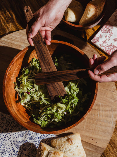 Overhead view of someone tossing a fresh green salad in a large wooden bowl using hand-carved wooden salad servers, with bread and napkins nearby.