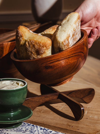 Hand reaching to serve freshly baked bread from a carved wooden bowl with curved stand, styled with a green mug and natural linen.