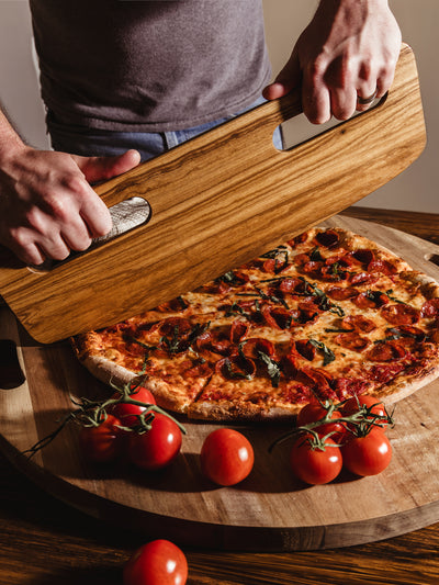 Close-up of hands using a light wooden pizza cutter to lift a freshly baked pepperoni pizza, with vine tomatoes in the foreground, celebrating the art of handcrafted kitchenware.