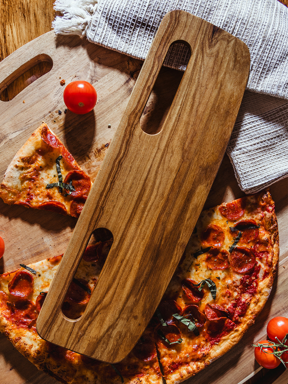A light wooden pizza cutter resting on a pizza-laden cutting board, surrounded by fresh tomatoes and textured kitchen towels, capturing the warmth and simplicity of artisan dining.