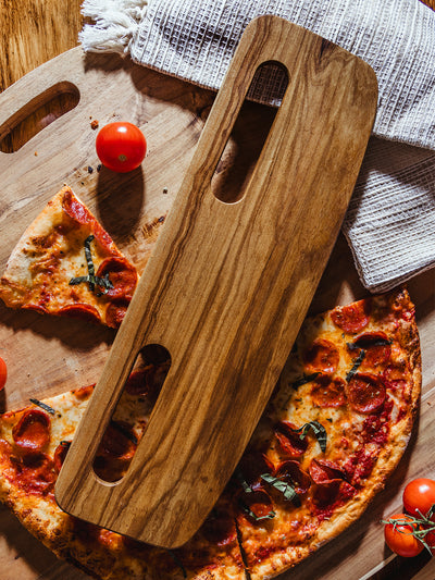 A light wooden pizza cutter resting on a pizza-laden cutting board, surrounded by fresh tomatoes and textured kitchen towels, capturing the warmth and simplicity of artisan dining.
