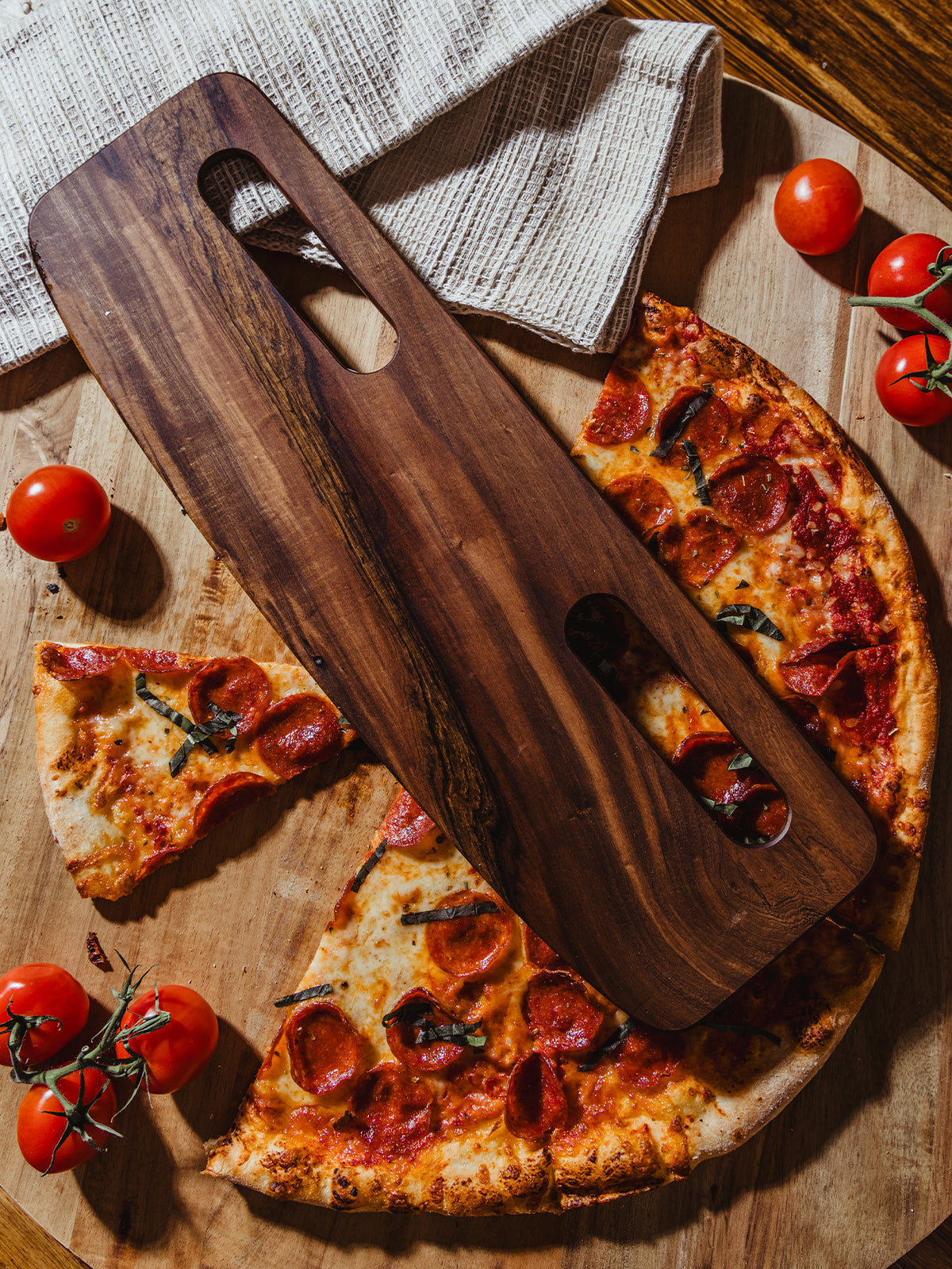 A dark wooden pizza cutter with a prominent grain pattern, positioned over slices of pepperoni pizza, framed by fresh vine tomatoes and a natural wooden backdrop for a rustic, handcrafted feel.