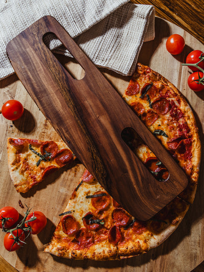 A dark wooden pizza cutter with a prominent grain pattern, positioned over slices of pepperoni pizza, framed by fresh vine tomatoes and a natural wooden backdrop for a rustic, handcrafted feel.