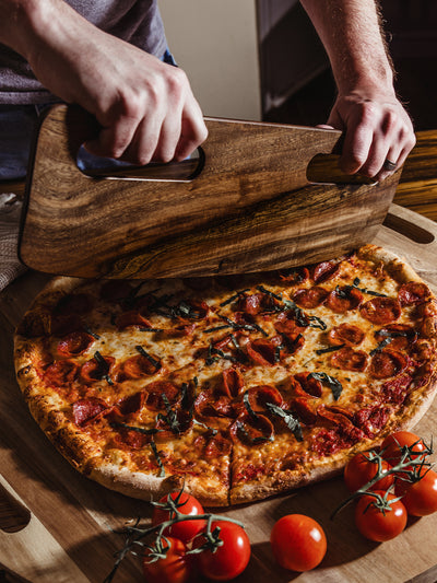 Hands gripping the smooth cutouts of a handcrafted wooden pizza cutter, lifting a full pepperoni pizza, emphasizing the cutter’s sturdy construction and artisan quality.
