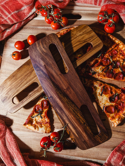 Handcrafted wooden pizza cutters layered over a pepperoni pizza, surrounded by fresh vine tomatoes and rustic red linens, showcasing the natural wood grain and ergonomic design.