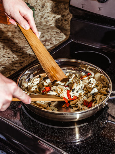 Hands using handcrafted wooden cooking utensils to stir a vibrant pasta dish with roasted red peppers and herbs in a stainless steel skillet, capturing the warmth and functionality of artisan kitchen tools.