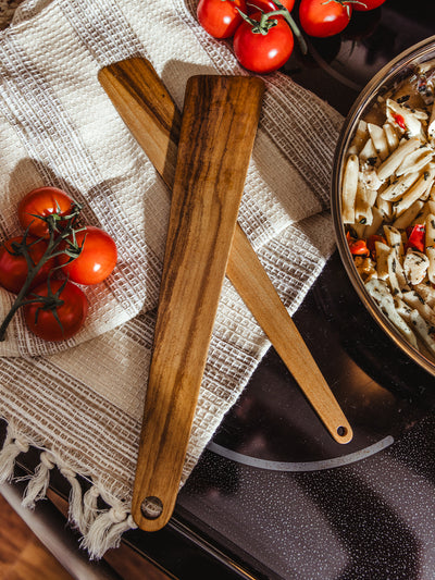 Two handcrafted wooden cooking spatulas resting on a textured cotton kitchen towel, alongside vine-ripened tomatoes and a pan of pasta, highlighting the natural grain and clean design.