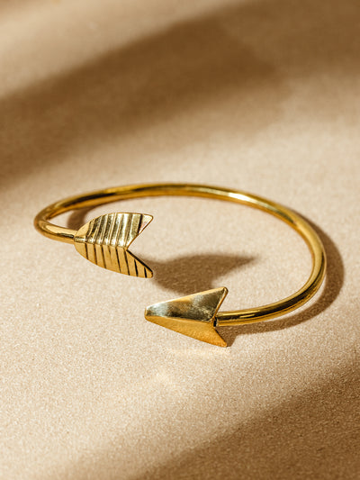 Gold-toned brass cuff bracelet featuring an open-ended arrowhead design, displayed on a warm sand-colored background. One end of the cuff has a smooth arrowhead, while the other features a textured, lined detail, adding a rustic and handcrafted aesthetic.