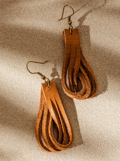Pair of handcrafted looped leather earrings with brass hooks, laid on a neutral textured surface in warm natural light.