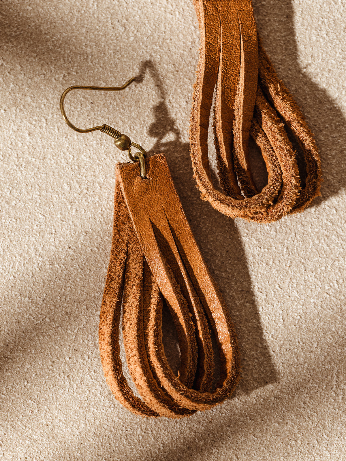 Close-up of a single looped leather earring with brass hook, showing the raw and smooth leather textures on a neutral surface.