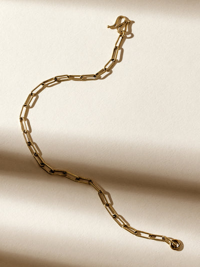 Gold chain link bracelet with a hook clasp, photographed on a textured cream background with soft natural shadows, highlighting its minimalist and elegant design.
