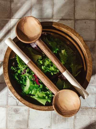 Set of two wooden salad servers with white carved handles resting on a bowl of fresh greens, placed on a rustic tiled surface.