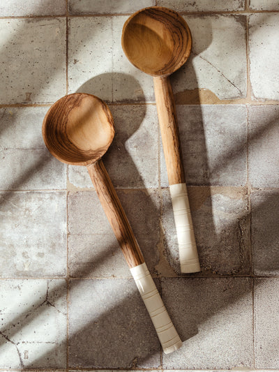 Pair of handcrafted wooden serving spoons with white carved handles, styled on rustic tile in warm natural light.
