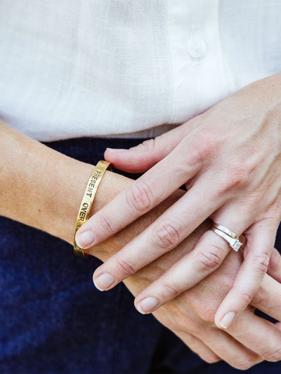 Close-up of a woman's hands wearing a gold cuff bracelet engraved with the phrase "PRESENT OVER PERFECT" and a silver ring set.