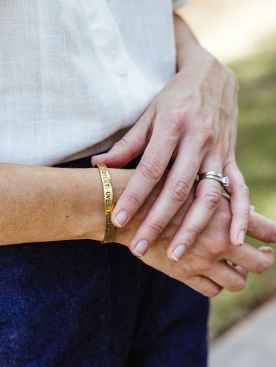 Woman's wrist adorned with a gold engraved cuff bracelet that reads "PRESENT OVER PERFECT," styled with a silver engagement and wedding ring.
