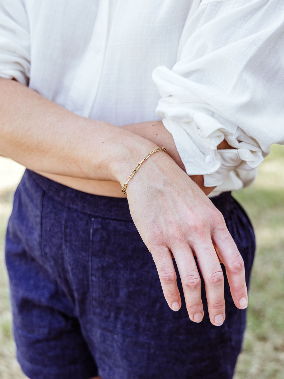Woman wearing a delicate gold chain link bracelet with rolled sleeves and navy shorts, standing outdoors.