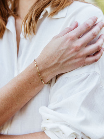 Close-up of a woman’s wrist wearing a minimal gold chain link bracelet over a rolled white sleeve blouse.