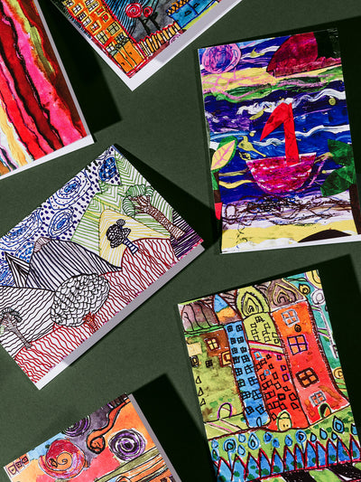 Colorful greeting cards laid out on a green surface, featuring vibrant, childlike illustrations including whimsical buildings, a swan on water, swirling skies, and abstract patterns.
