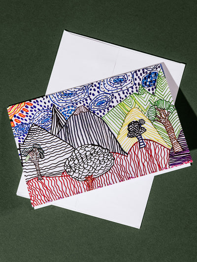 A greeting card resting on a white envelope, decorated with hand-drawn mountains, trees, and swirling sky patterns in red, blue, green, and black ink.