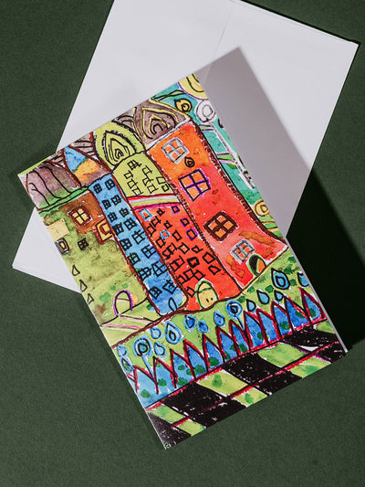 A vertically oriented greeting card on a white envelope featuring a bright, abstract drawing of tall, colorful buildings with whimsical windows and details.