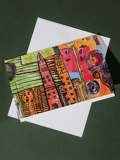 Horizontal greeting card showing a vivid, artistic interpretation of tall, colorful buildings, faces, and swirling shapes, paired with a white envelope.