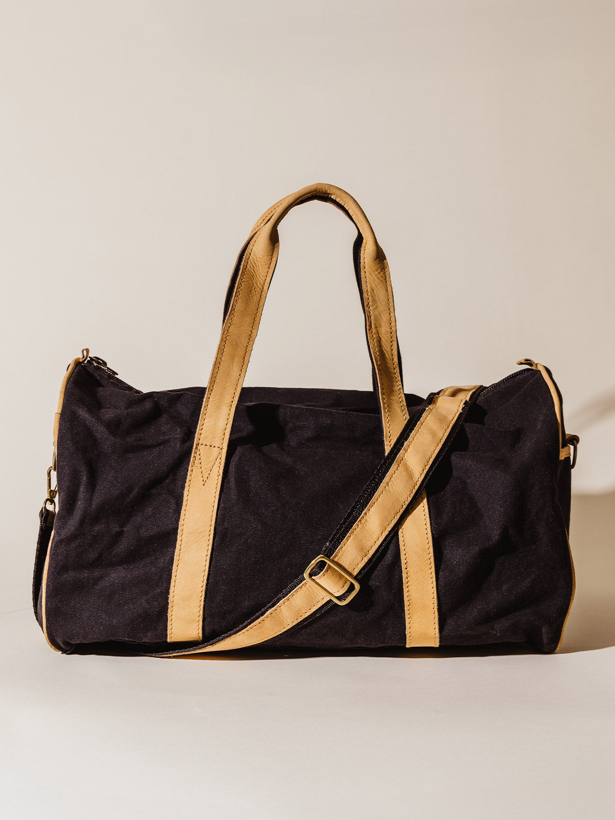 Black canvas duffel bag with tan leather handles and shoulder strap, shown upright against a neutral background.