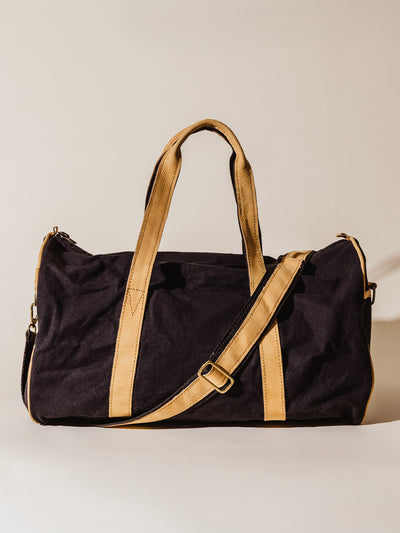 Black canvas duffel bag with tan leather handles and shoulder strap, shown upright against a neutral background.