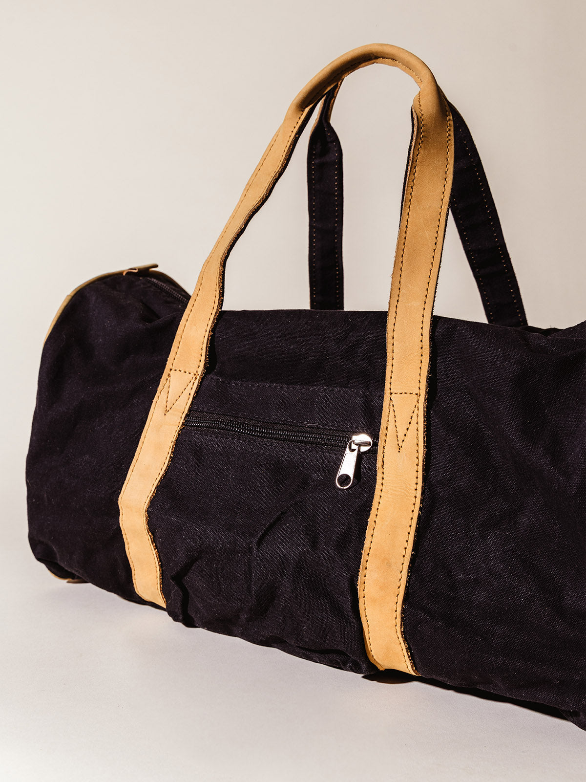 Back view of a black canvas duffel bag featuring a zippered pocket and tan leather handles, photographed in soft studio lighting.